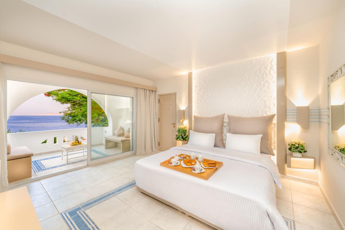 Une chambre d’hôtel avec un grand lit, un balcon donnant sur la mer et une table à manger avec des produits pour le petit-déjeuner.