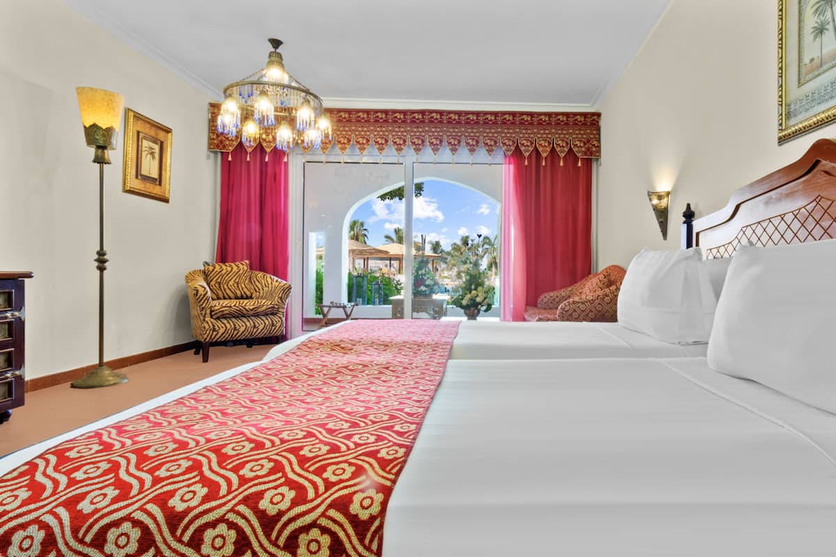 Une chambre d’hôtel avec deux lits, une moquette rouge à motifs, un fauteuil à rayures de tigre, un lustre et une vue sur une piscine et des palmiers.