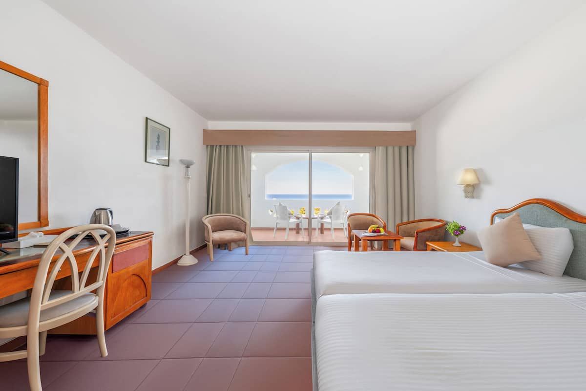 Une chambre d’hôtel avec un lit, un bureau et une chaise, une petite table avec une lampe, et une vue sur la mer par une grande fenêtre.
