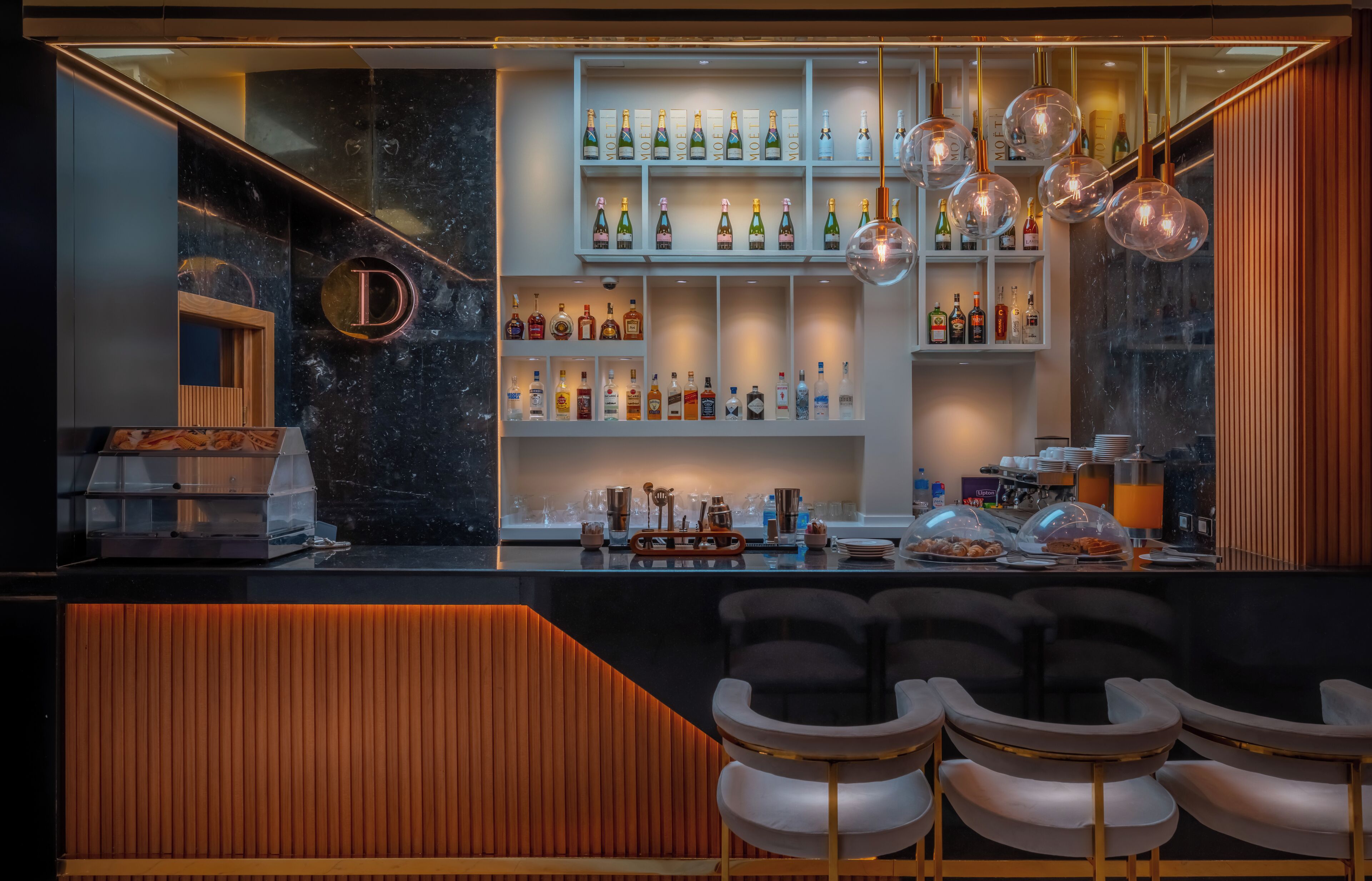 Zona de bar moderna con encimera de mármol, botellas de licores y luminarias colgantes.