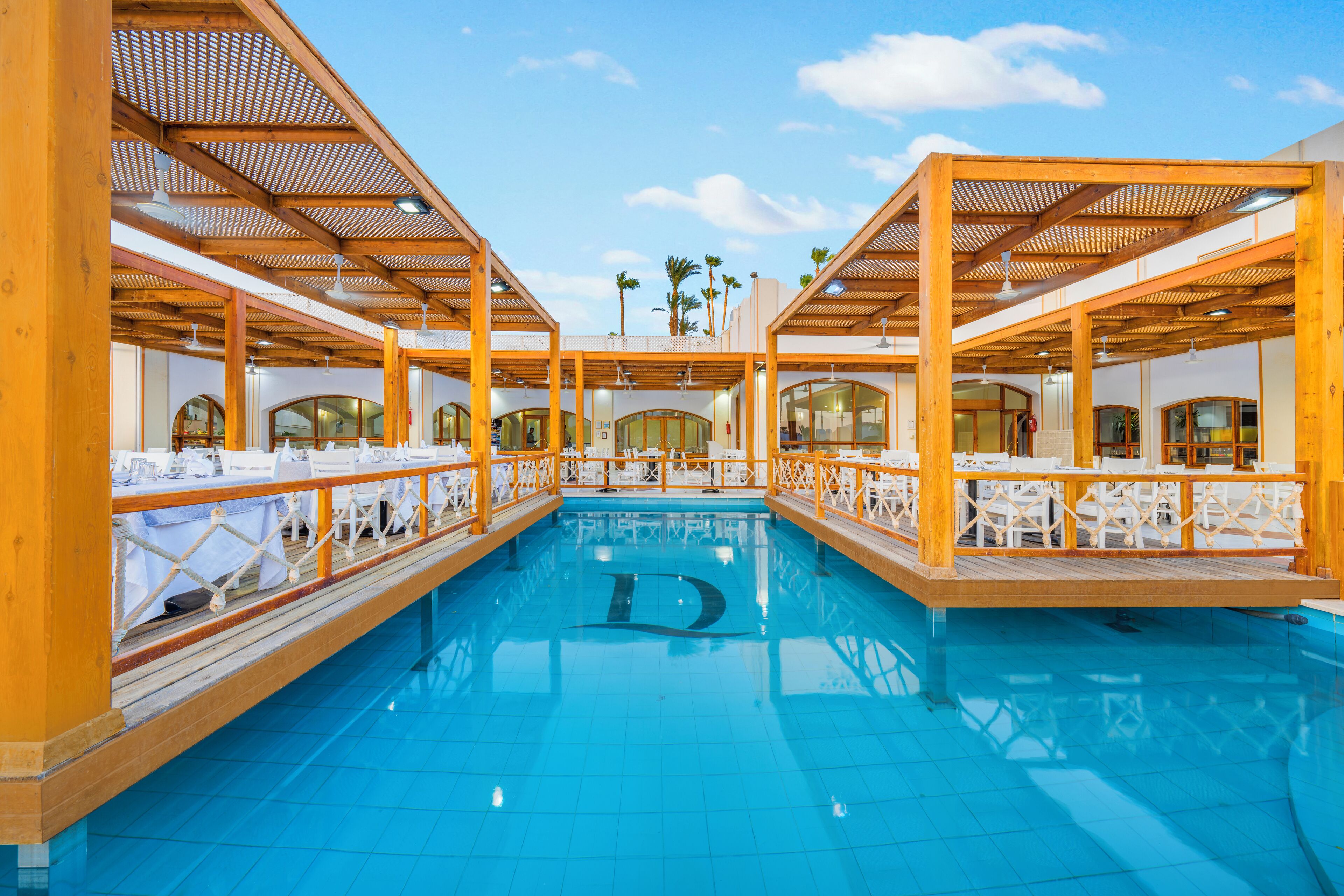 A swimming pool with wooden deck seating and tables, surrounded by a covered area with a view of palm trees.