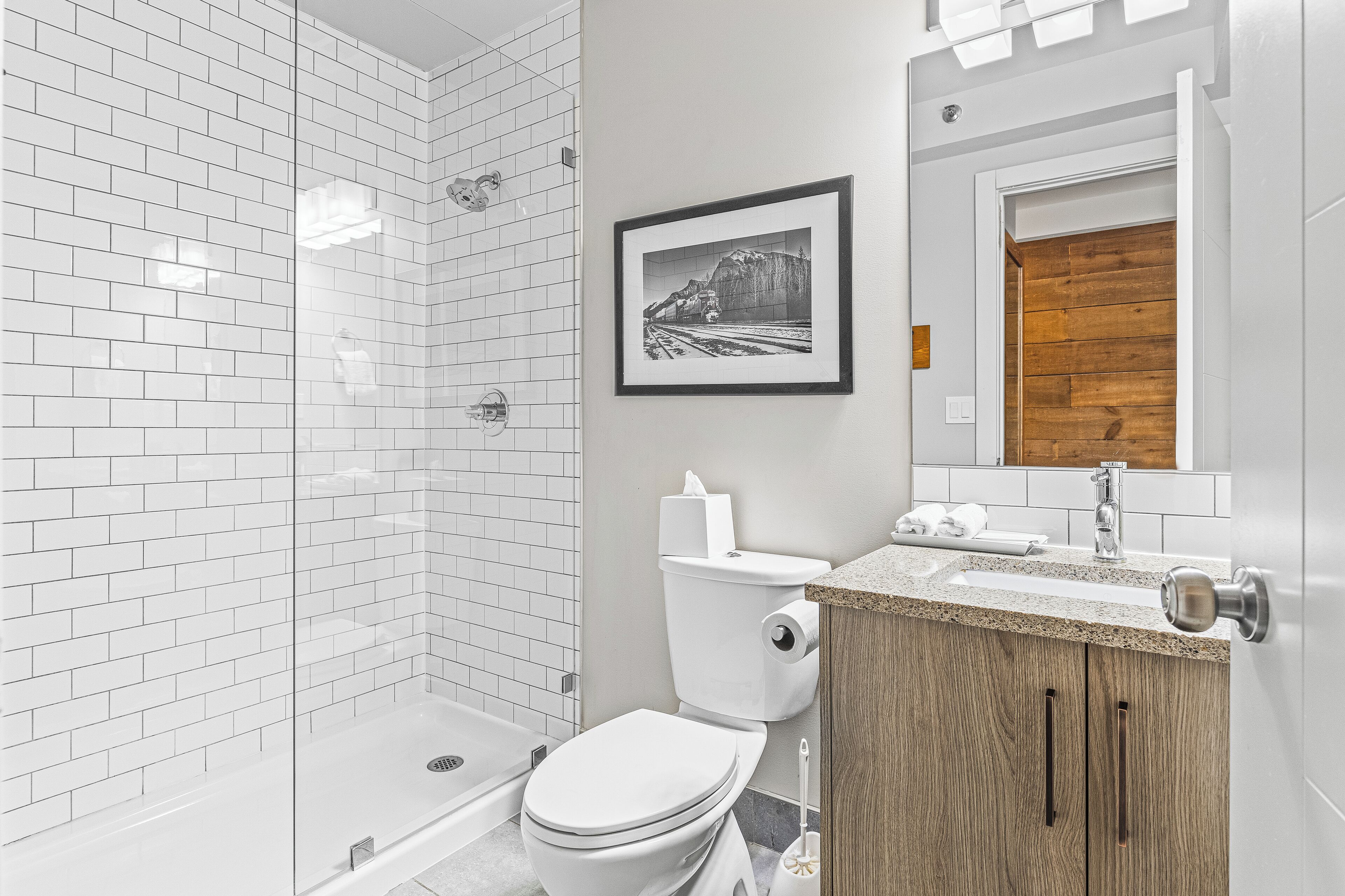 A modern bathroom with white subway tiles, a glass shower enclosure, a toilet, and a wooden vanity with a sink.