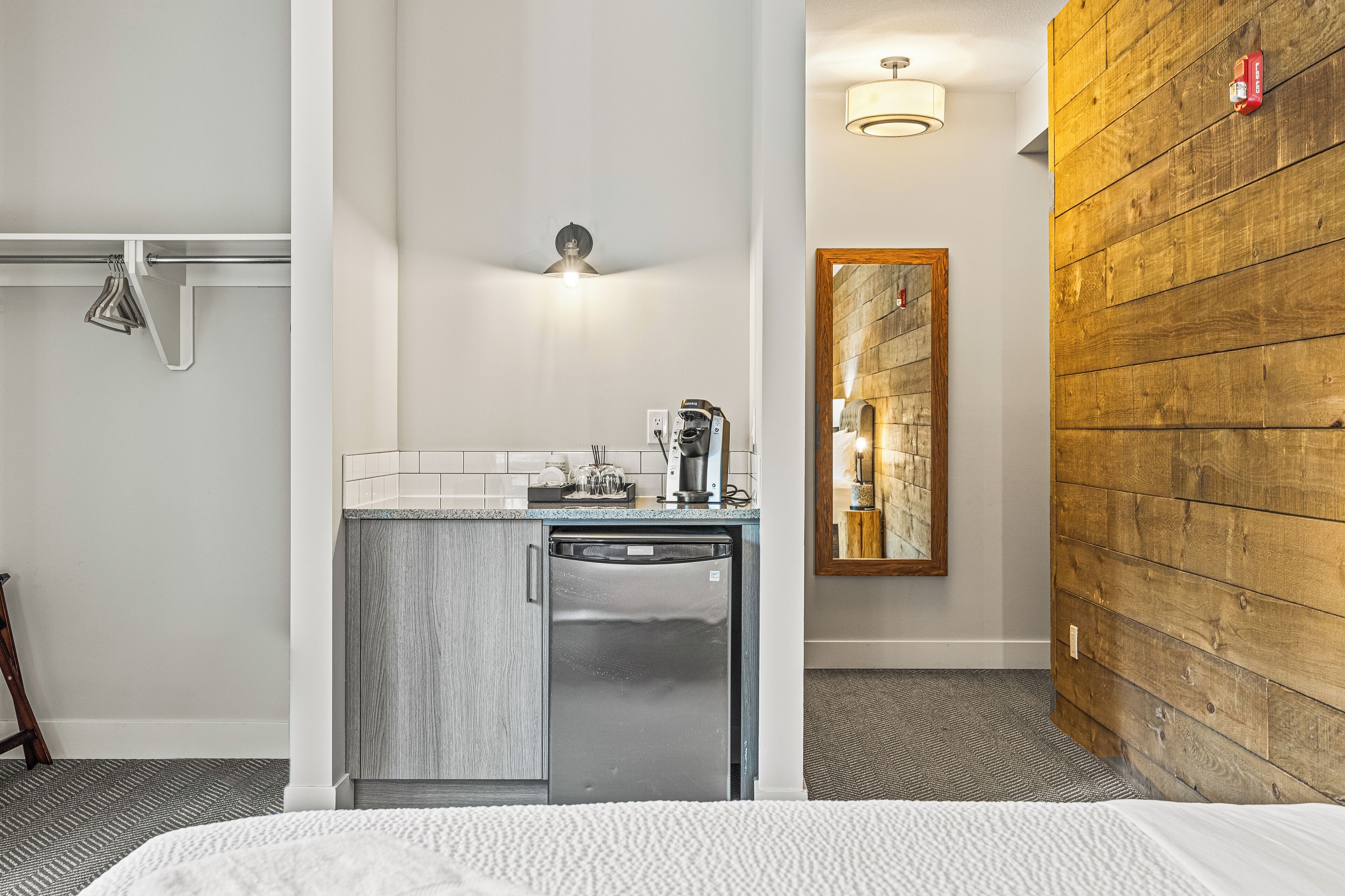 A compact hotel room with a kitchenette, a small sink, a mirror, and a wooden accent wall.