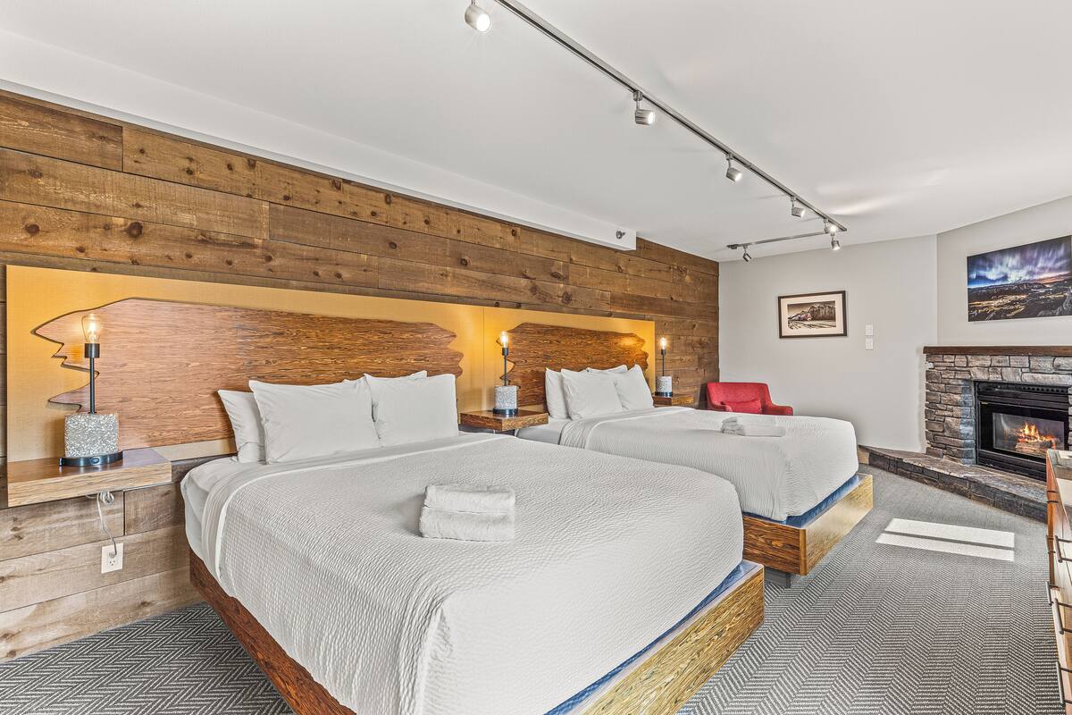 A modern hotel room with two beds, a fireplace, and a wooden accent wall.