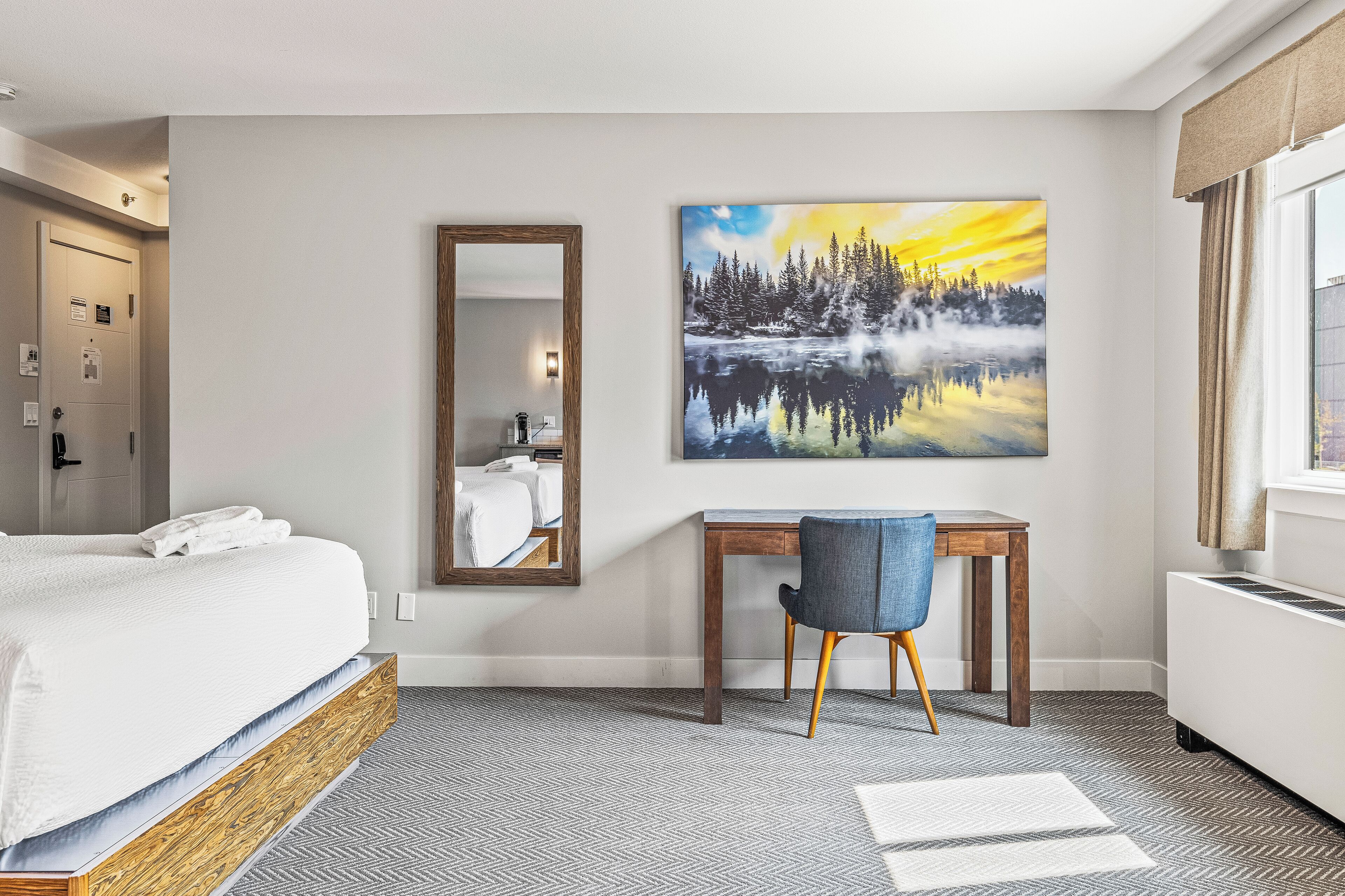 Une chambre d’hôtel comprenant un lit, un bureau, une chaise, un grand miroir et un tableau représentant une forêt et un lac.