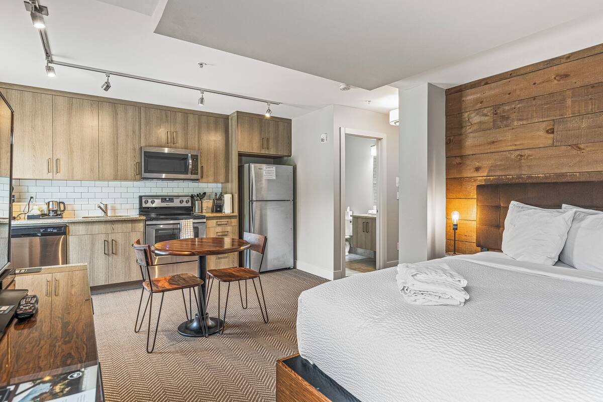 A modern hotel room with a large bed, a wooden headboard, a kitchenette with a microwave and refrigerator, a dining area with a table and chairs, and a bathroom with a mirror and sink.