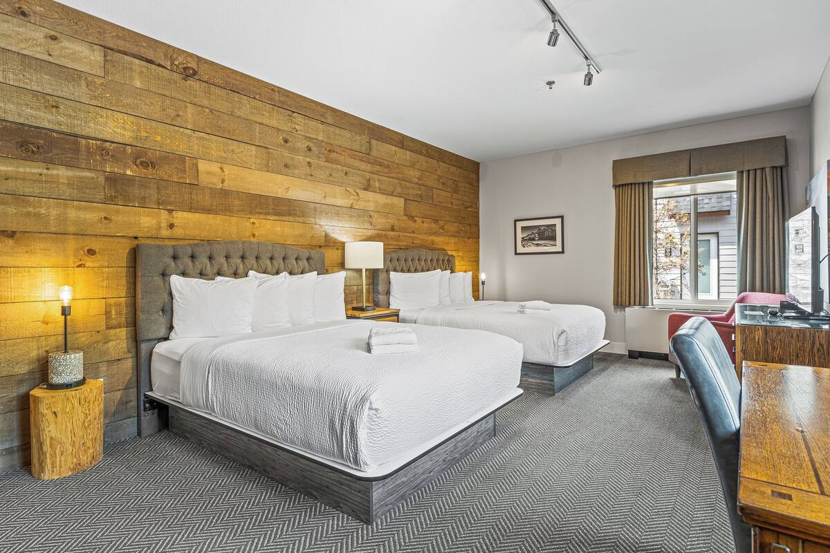 A hotel room with two beds, wooden panel walls, a herringbone carpet, a wooden nightstand, and a desk with a chair.