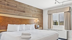 A bedroom with a wooden headboard, a large bed with white bedding, a bedside table with a lamp, and a window with curtains. - Basecamp Lodge Canmore (Canmore)