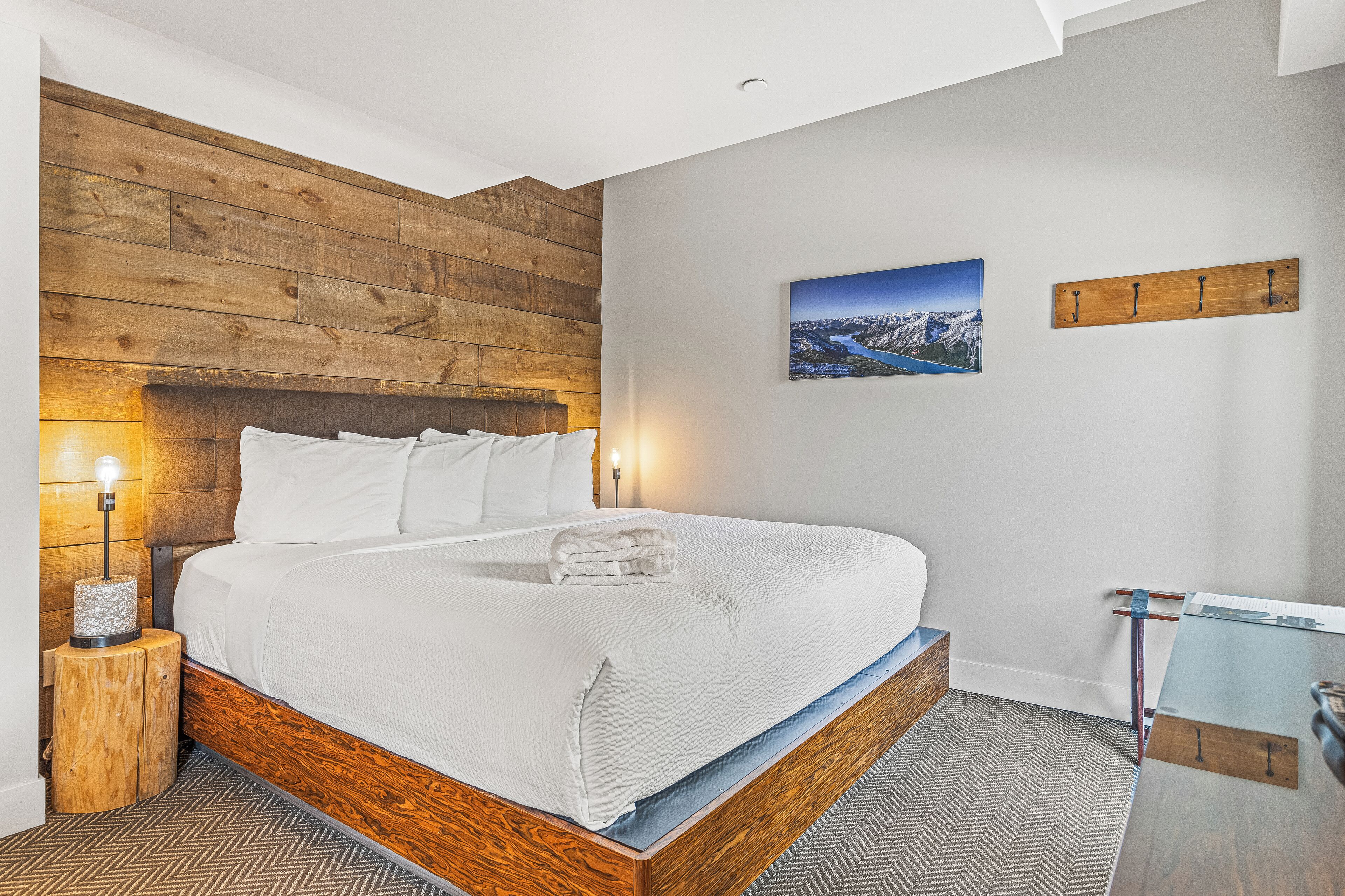 Un dormitorio moderno con cabecera de madera, cama bien hecha, mesita de noche con lámpara y perchero en la pared.