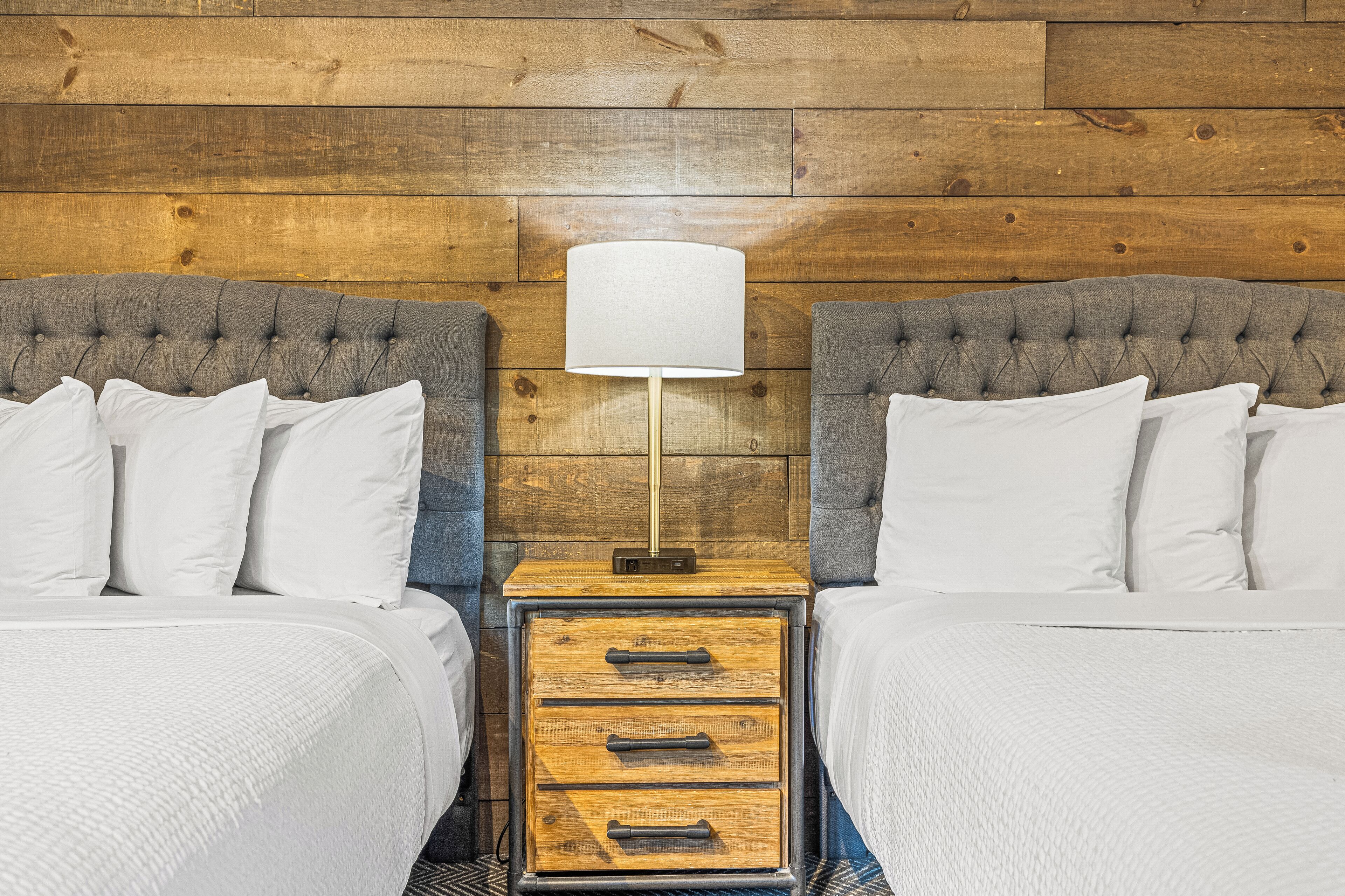 A double bed with a grey headboard, a bedside table with a lamp, and a wooden wall paneling.