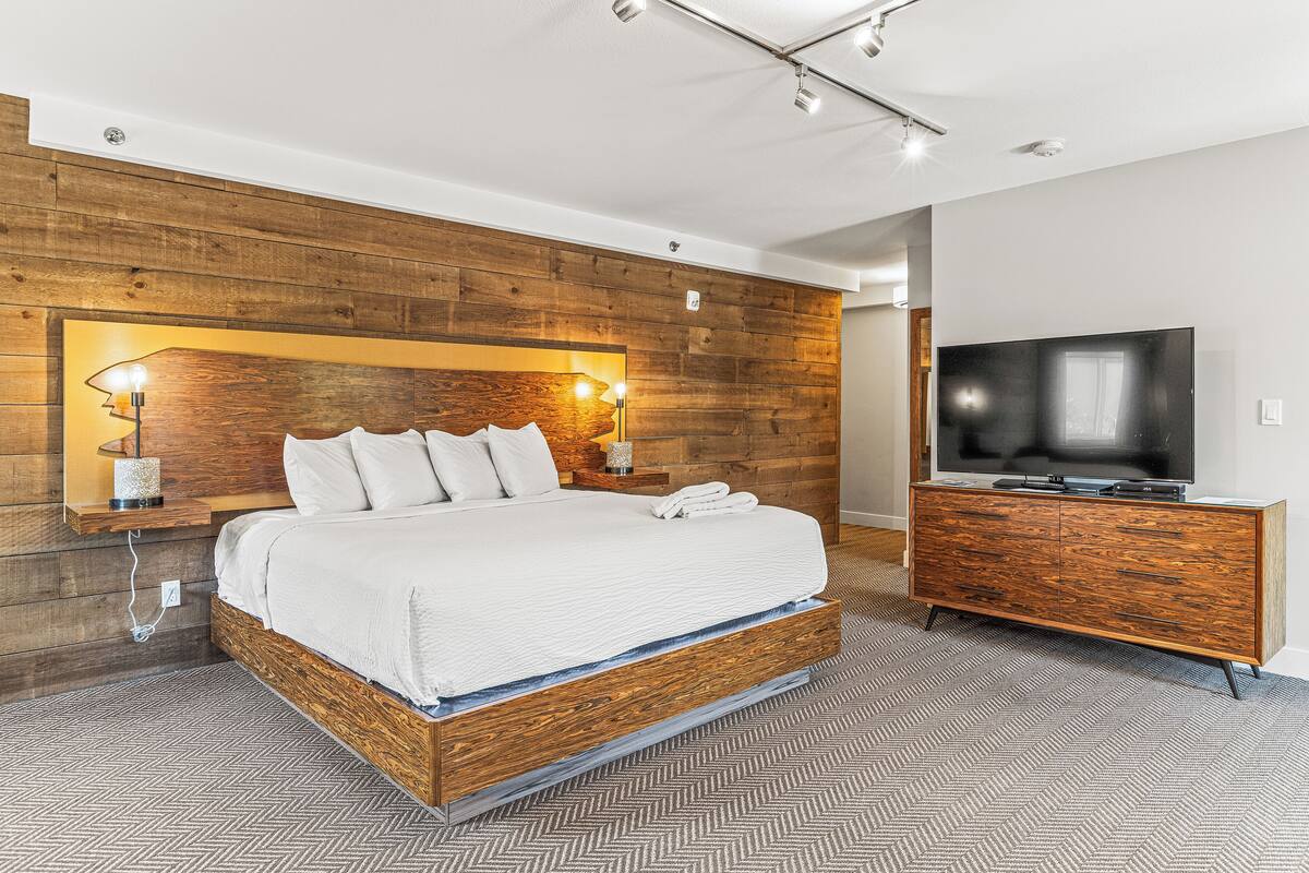 A modern hotel room with a wooden headboard, a large bed, a TV, and a wooden dresser.
