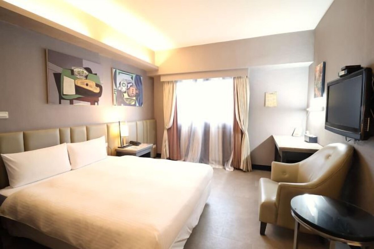 A hotel room with a large bed, a desk with a chair, a television, and artwork on the walls.