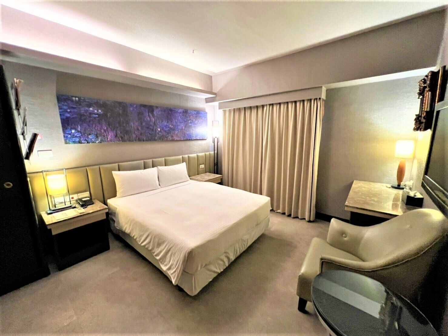 A hotel room with a large bed, a desk with a lamp, a chair, and a large window with curtains.
