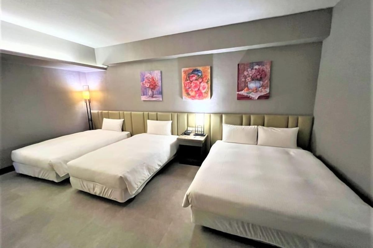 Habitación de hotel con dos camas individuales y una cama doble, una mesita de noche con lámpara, y tres cuadros con motivos florales en la pared.