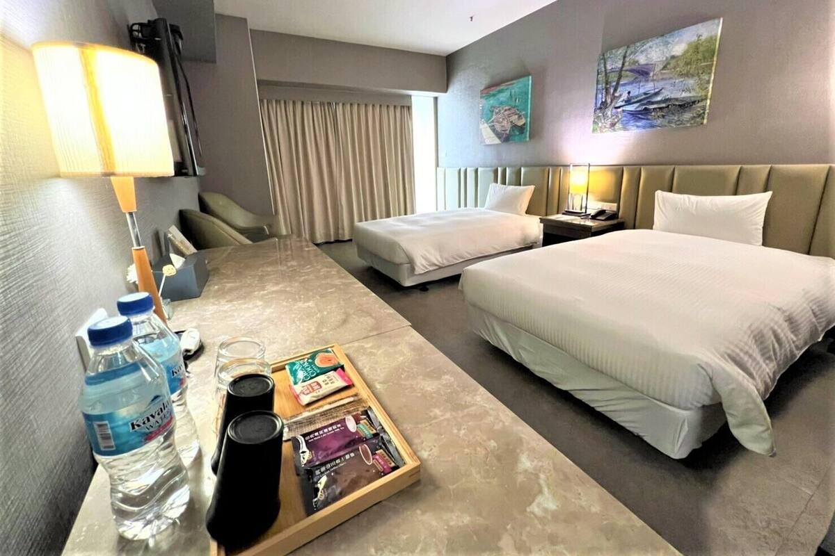 Une chambre d’hôtel avec un lit, un bureau avec une lampe, une chaise, une table de chevet et une fenêtre avec des rideaux. Il y a également deux tableaux accrochés au mur.