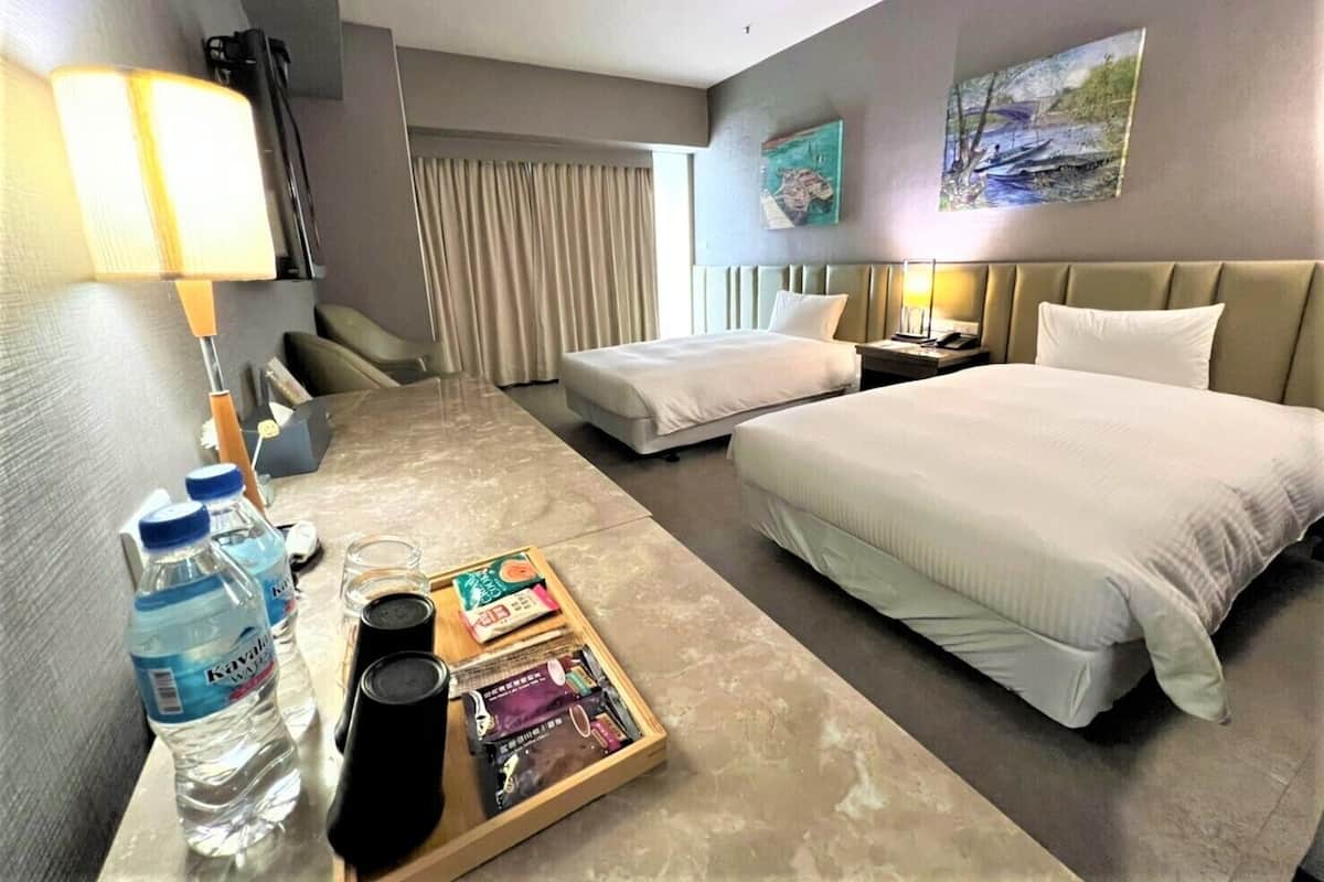 A hotel room with a bed, a desk with a lamp, a chair, a nightstand, and a window with curtains. There are also two paintings on the wall.