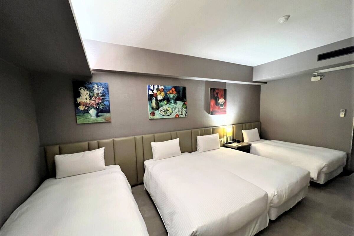 Une chambre d’hôtel avec trois lits simples, une tête de lit et une table de chevet avec une lampe.