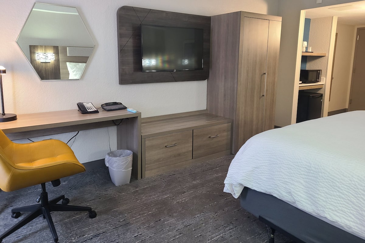 Ein Hotelzimmer mit einem Bett, einem Schreibtisch mit Stuhl, einem an der Wand befestigten Fernseher, einem Kleiderschrank und einem Mülleimer.