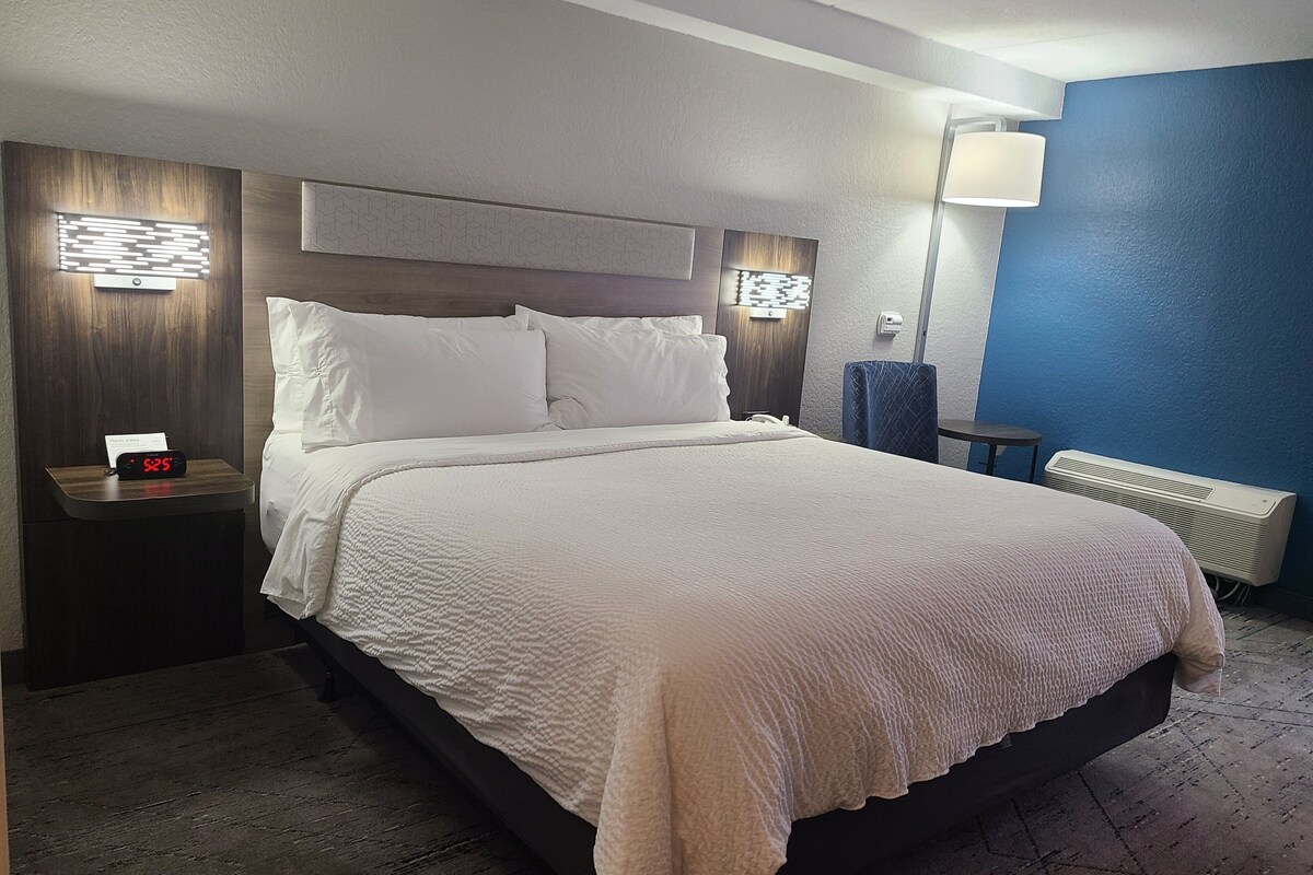 Una cama bien hecha con sábanas blancas, una mesita de noche con un reloj y una lámpara de pared.