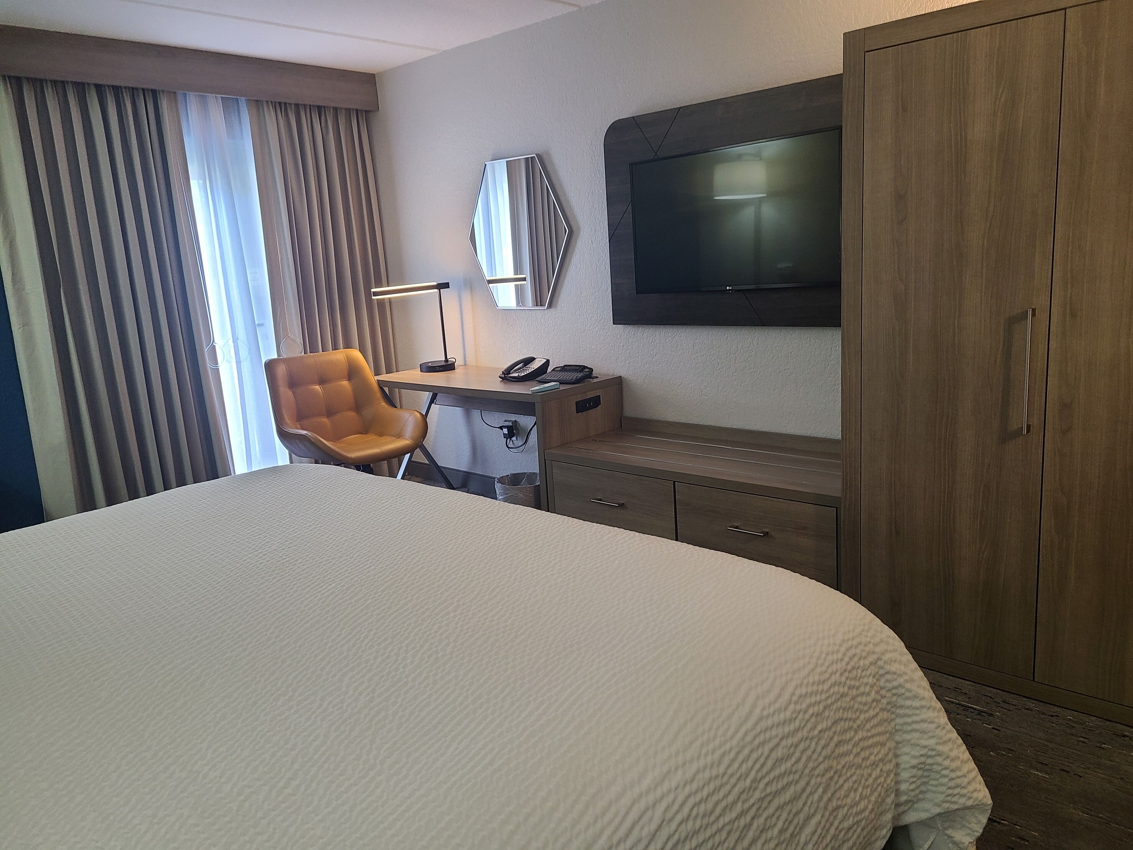 A hotel room with a large bed, a desk with a chair, a telephone, a mirror, and a television on the wall.