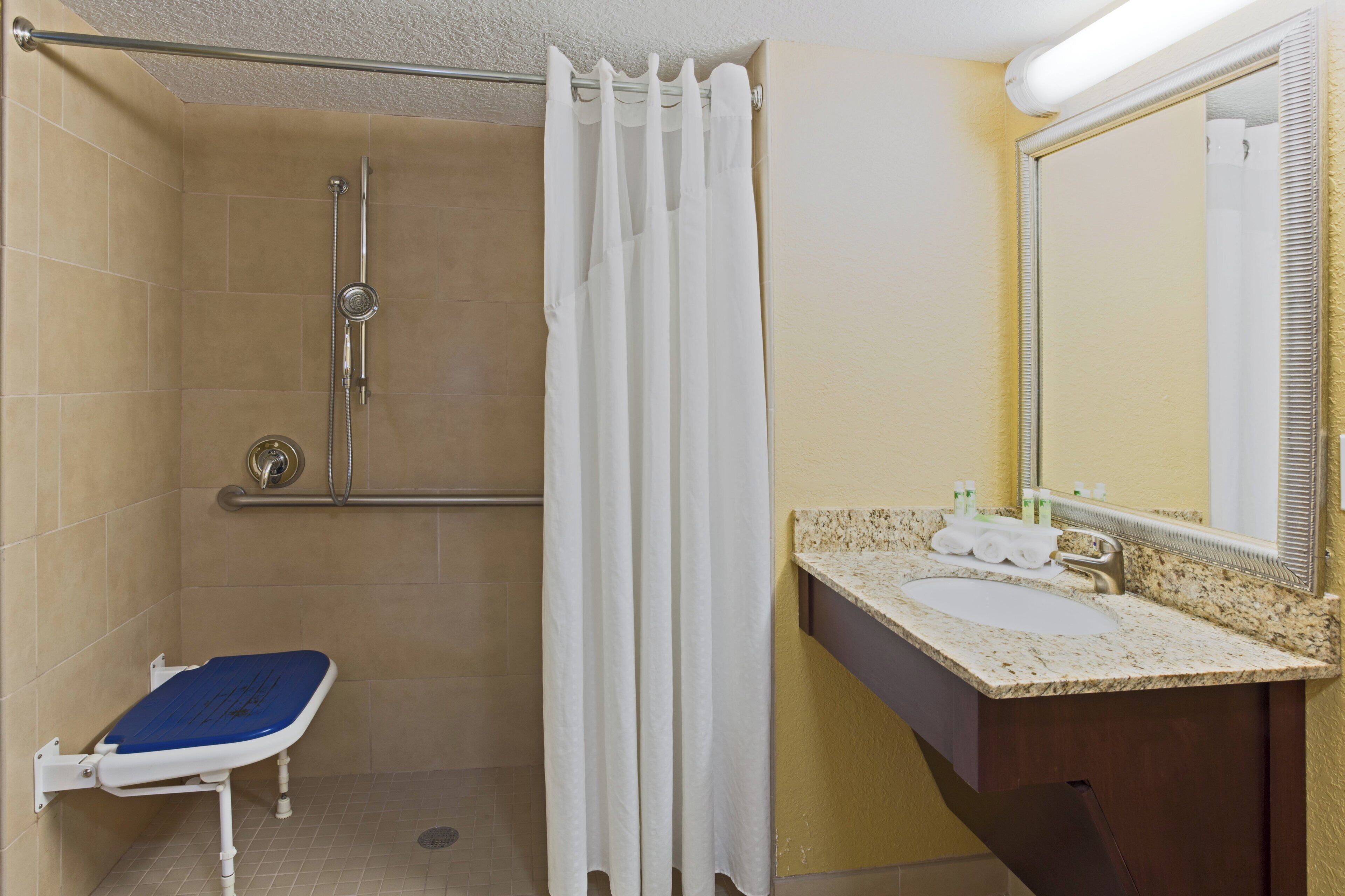 Un baño con ducha, inodoro con asiento azul, lavabo con espejo y toallero.