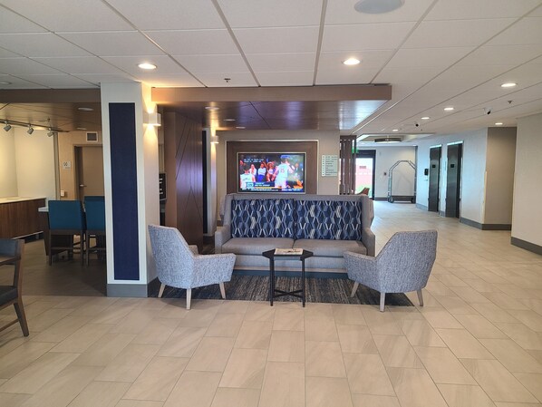 A lobby with a sofa, two armchairs, a small table, and a TV screen displaying a sports event. - Holiday Inn Express & Suites Ft. Lauderdale N - Exec Airport by IHG (Fort Lauderdale)