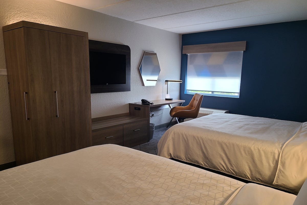 Ein Hotelzimmer mit zwei Betten, einem Schreibtisch, einem Stuhl, einem Kleiderschrank, einem Fernseher und einem Fenster mit Jalousien.