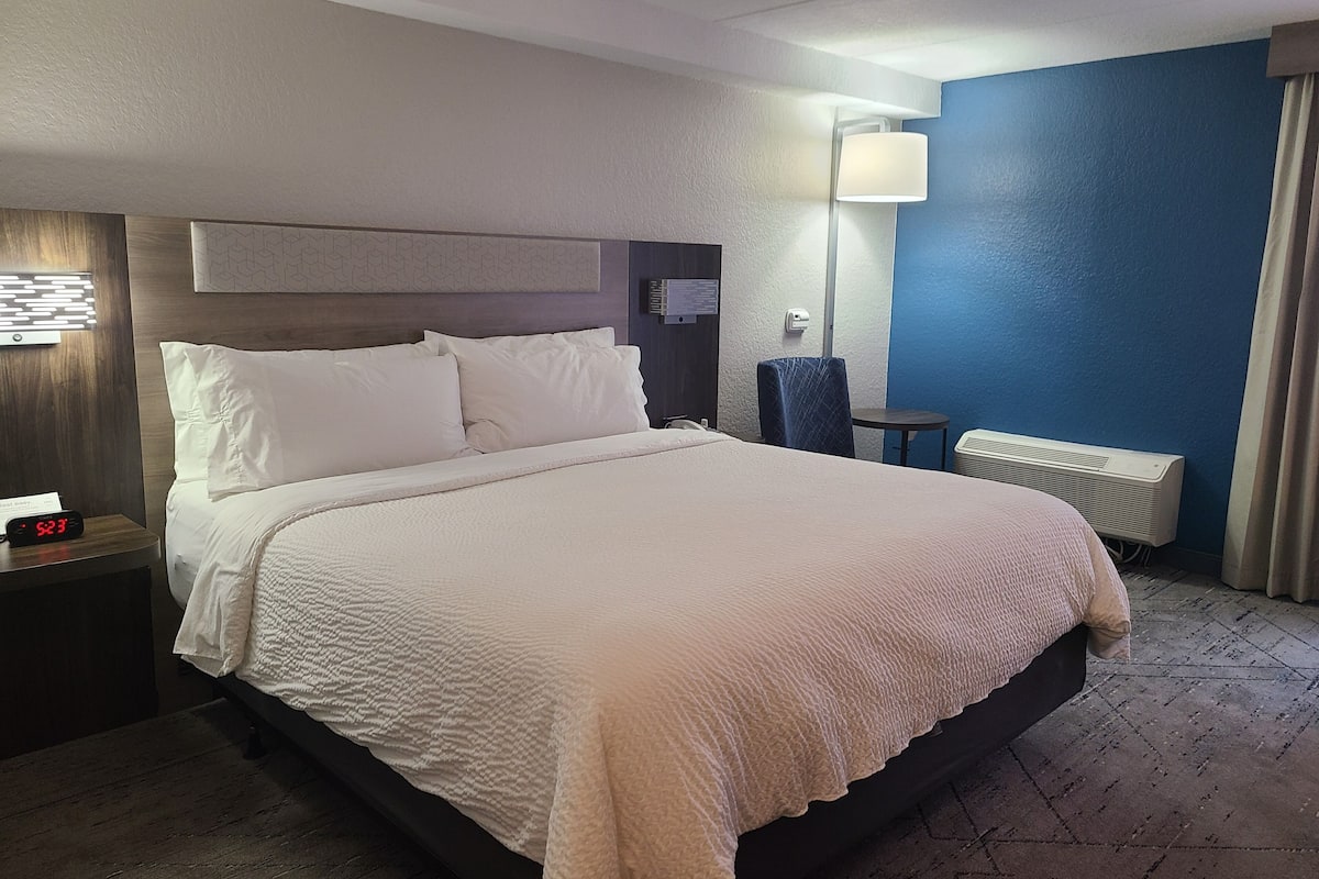 Ein Hotelzimmer mit einem großen Bett, einem Nachttisch mit einer Uhr, einer Schreibtischlampe, einem Stuhl und einem Heizkörper.