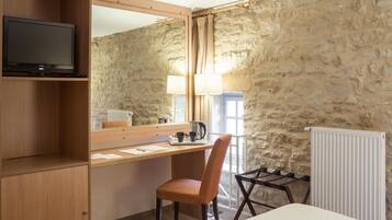 Una habitación de hotel con un escritorio de madera, una silla, una cama, un televisor en un estante y una pared de piedra.