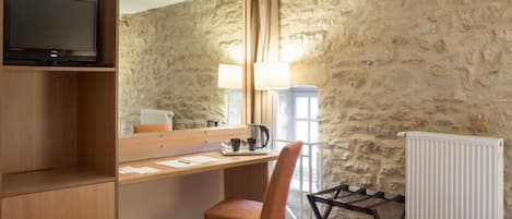 A hotel room with a wooden desk, a chair, a bed, a television on a shelf, and a stone wall.