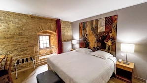A bedroom with a stone wall, a bed, a bench, a nightstand, and a painting. - Hotel Le Château Fort (Sedan)