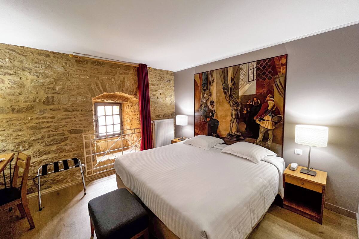 A bedroom with a stone wall, a bed, a bench, a nightstand, and a painting.
