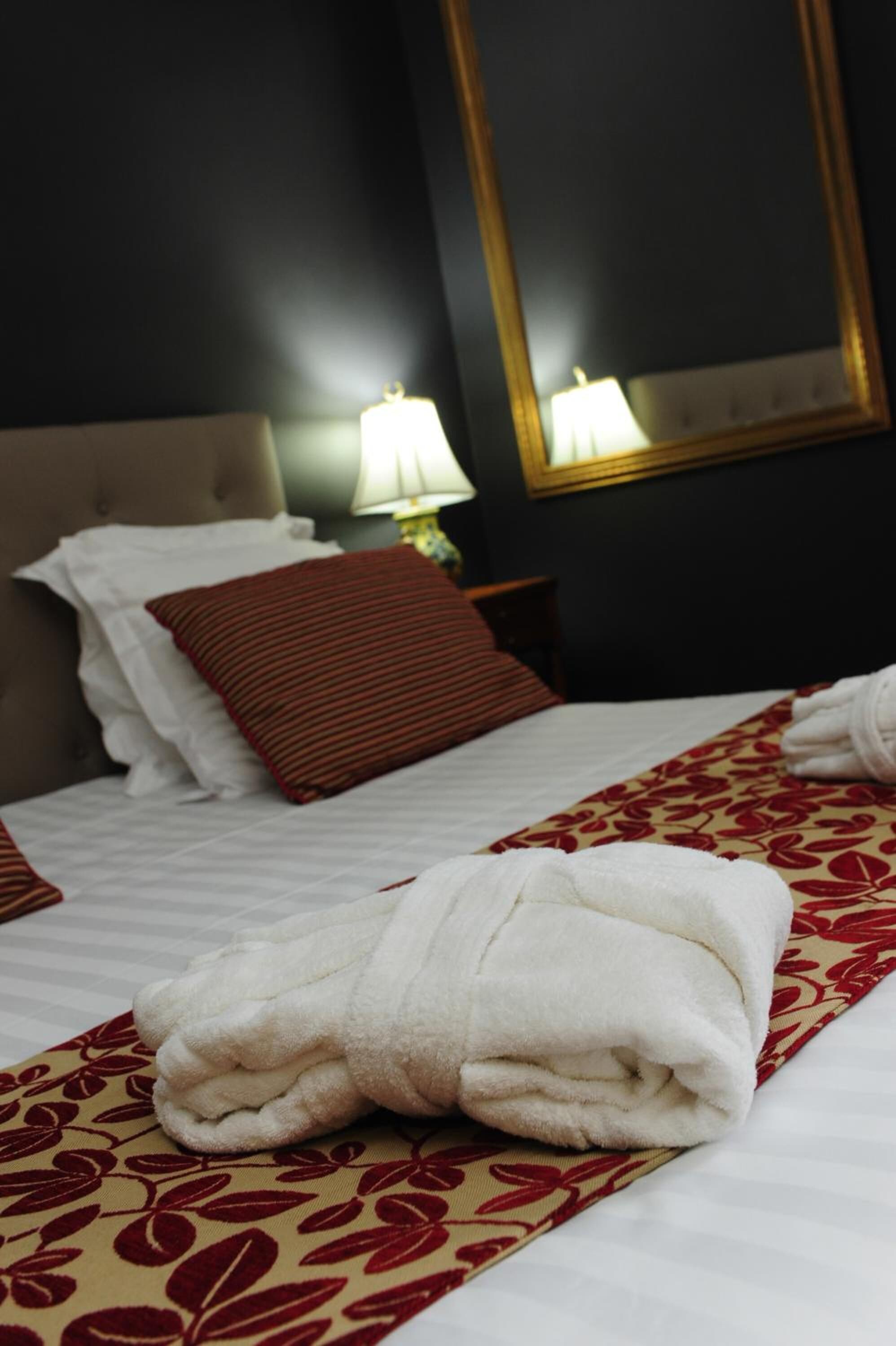 Cama bem arrumada com lençóis brancos, um cobertor estampado em vermelho e branco, e uma toalha branca dobrada.