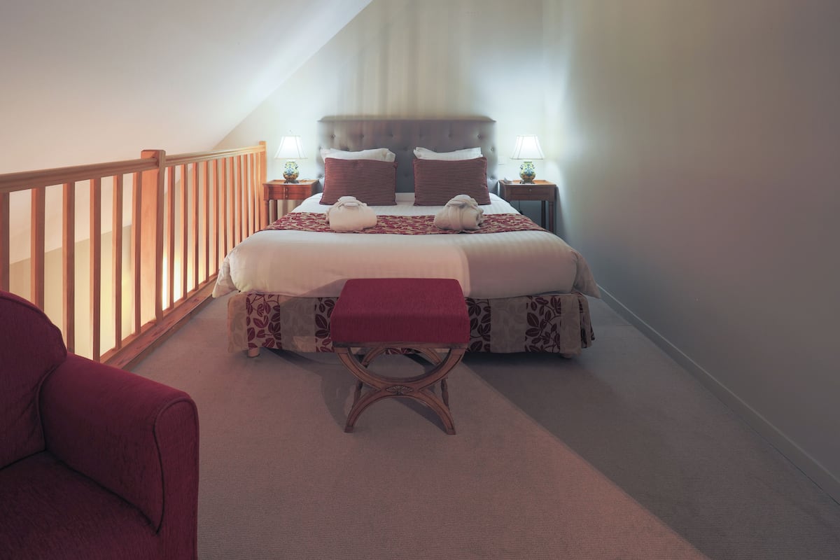 A bedroom with a bed, two bedside tables, a wooden bench, a red chair, and a wooden railing.