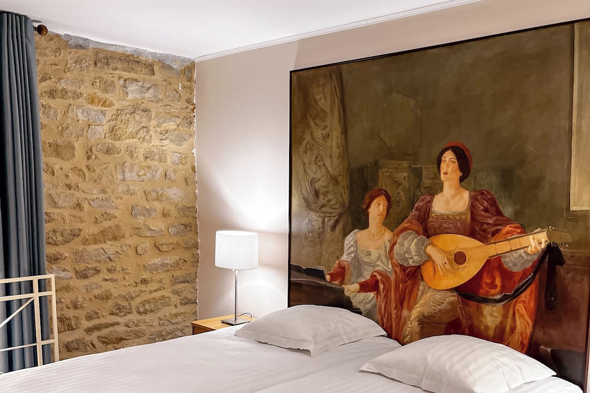 A double bed with white linens, a stone wall, and a large painting of two figures playing a guitar.