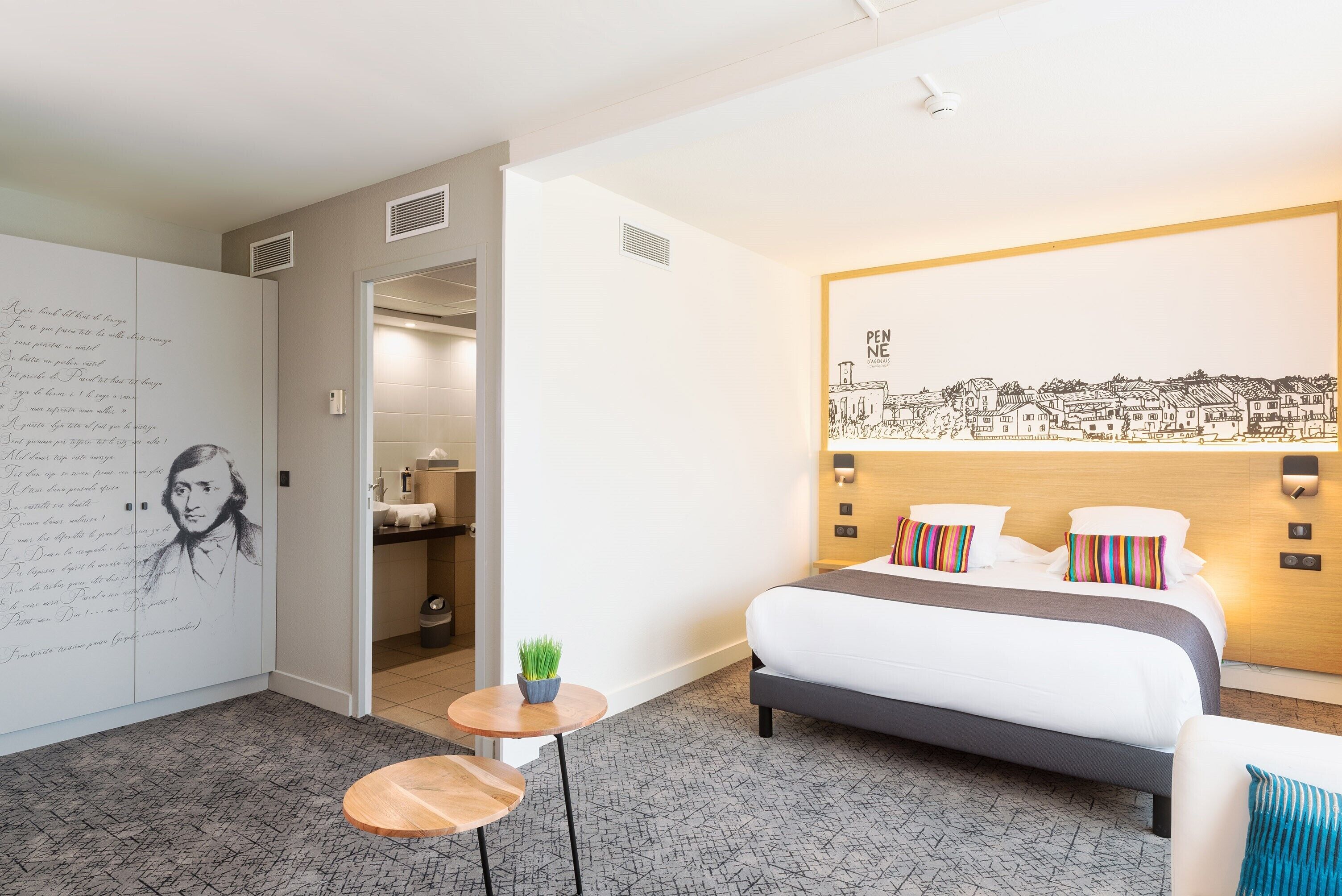 A modern hotel room with a large bed, a small round table with a plant, a wall with a portrait and a cityscape mural, and a bathroom visible through an open door.