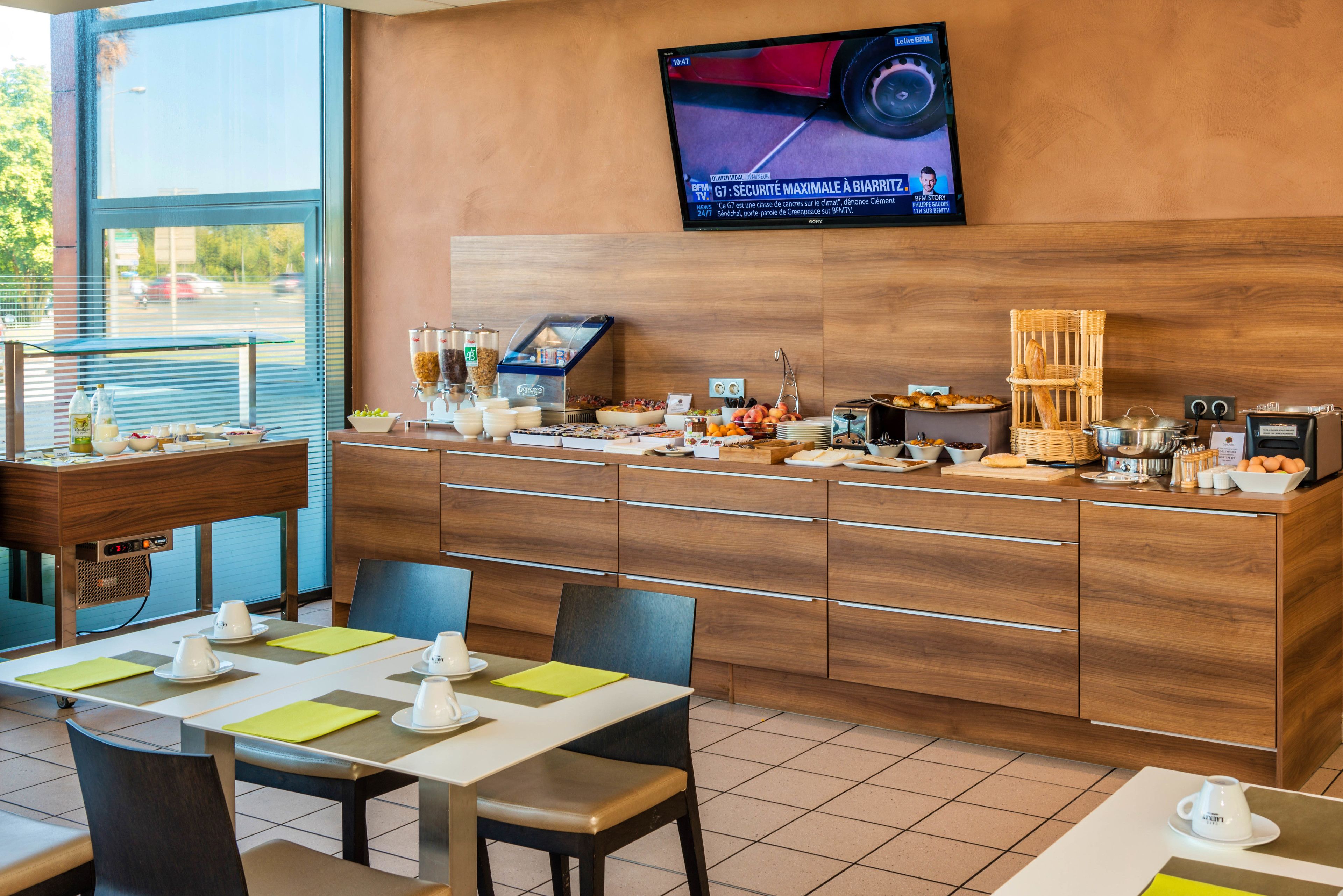 A breakfast area with a buffet setup, a television mounted on the wall, and a view of the outside through large windows.