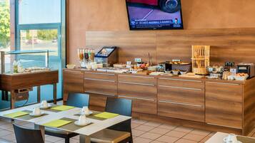 A breakfast area with a buffet setup, a television mounted on the wall, and a view of the outside through large windows.