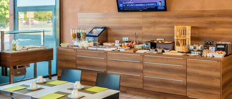 A breakfast area with a buffet setup, a television mounted on the wall, and a view of the outside through large windows.