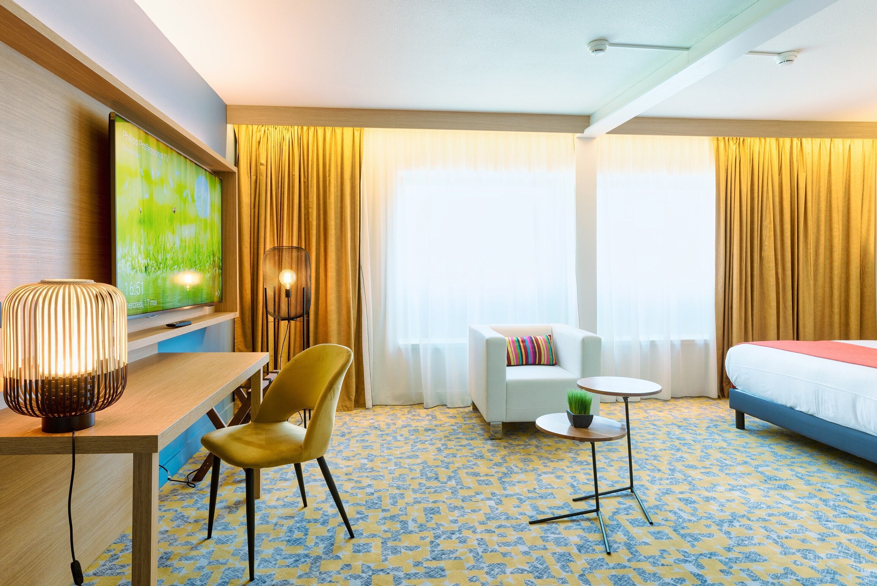 A hotel room with a bed, a desk with a lamp, a chair, a small table with a plant, and a TV on the wall.