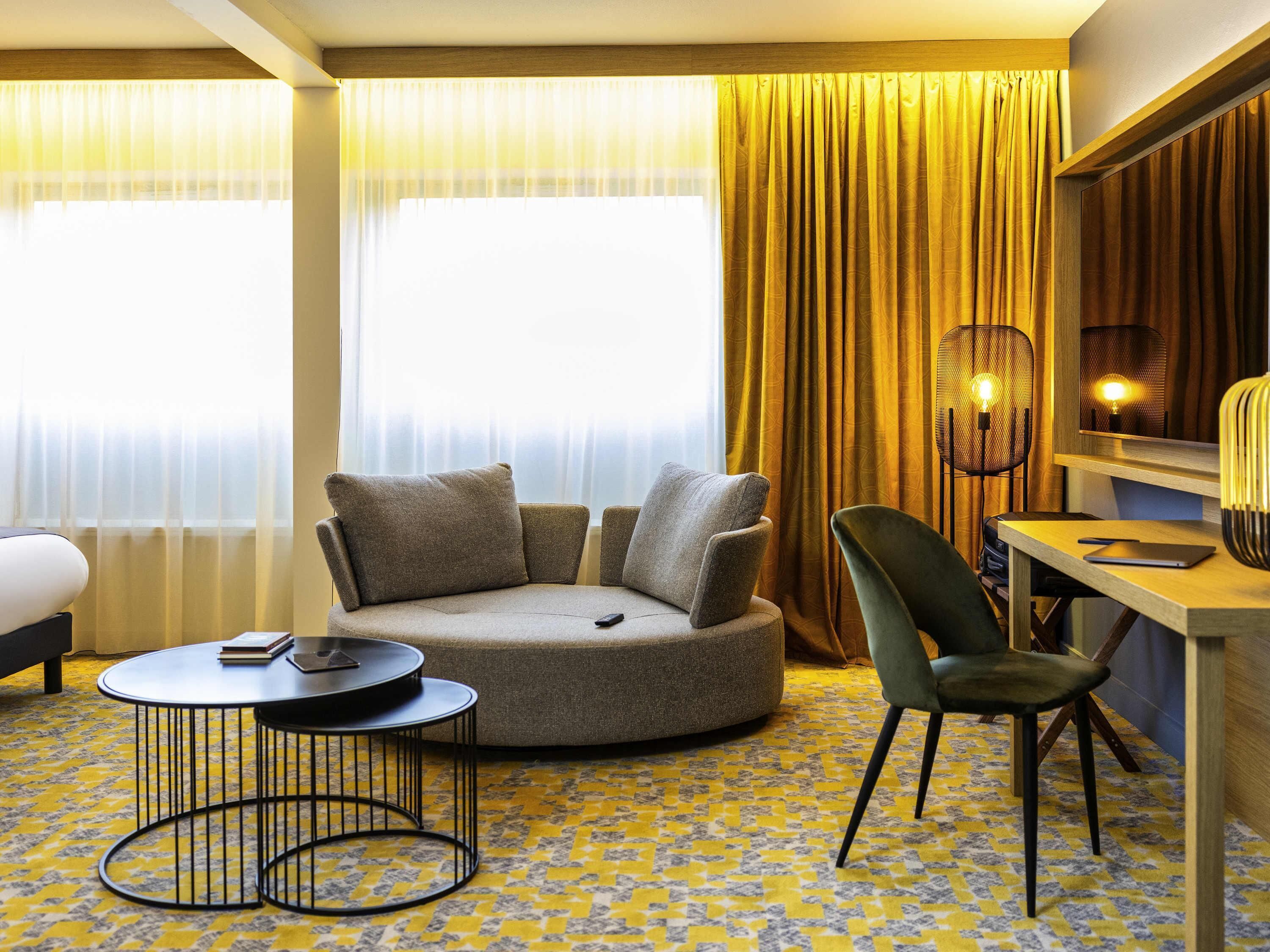 A hotel room with a round sofa, a desk, a chair, a small table, and a lamp.