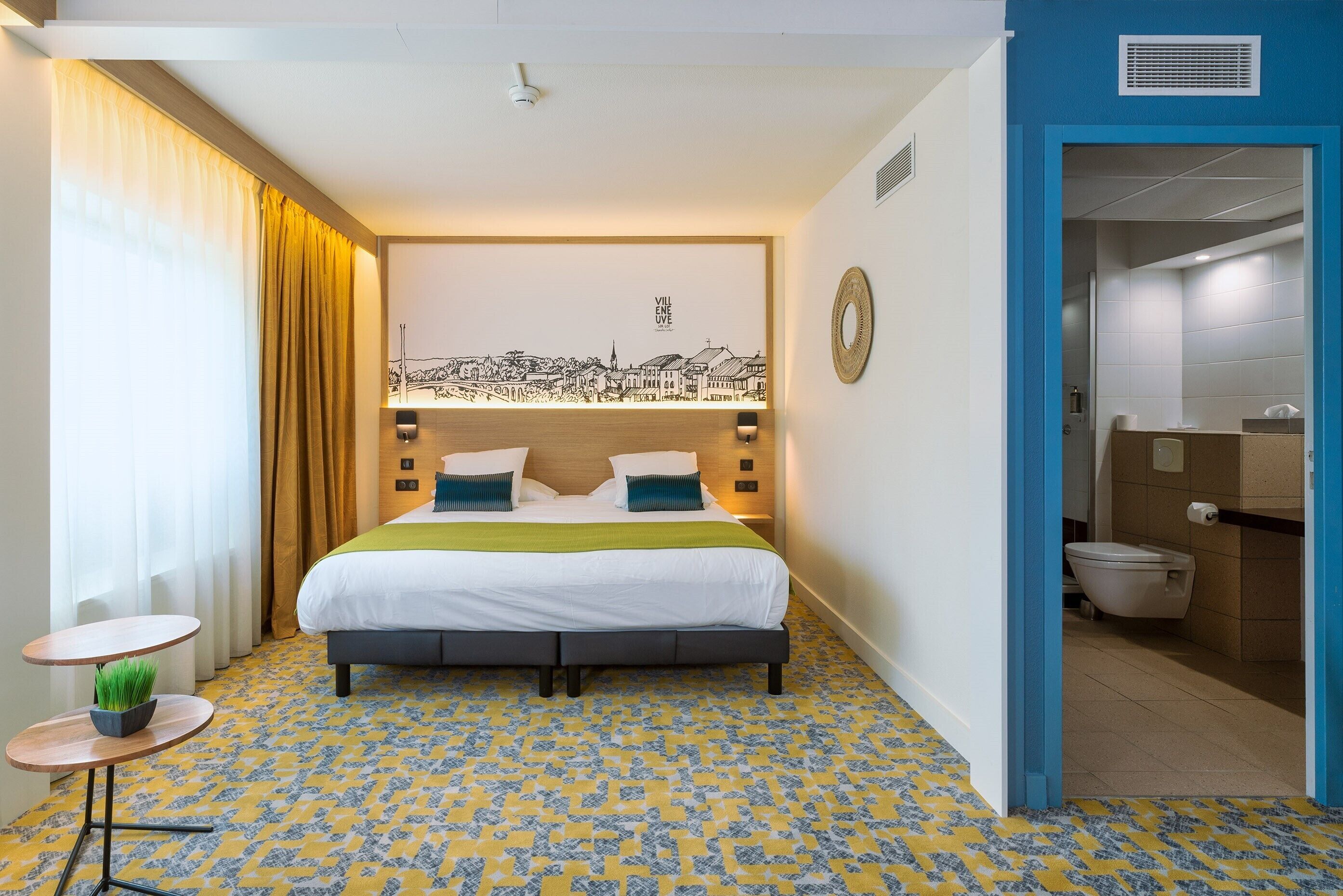 A hotel room with a bed, a small table with a plant, a cityscape artwork on the wall, and a bathroom visible through an open door.