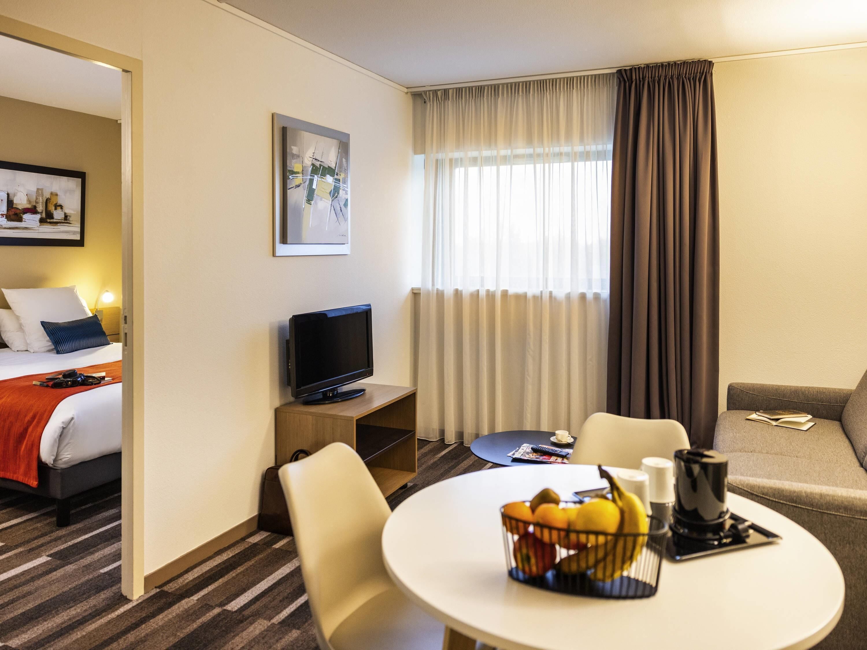 Una habitación de hotel con una cama, televisión, una mesa de comedor con fruta, un sofá y una ventana con cortinas.