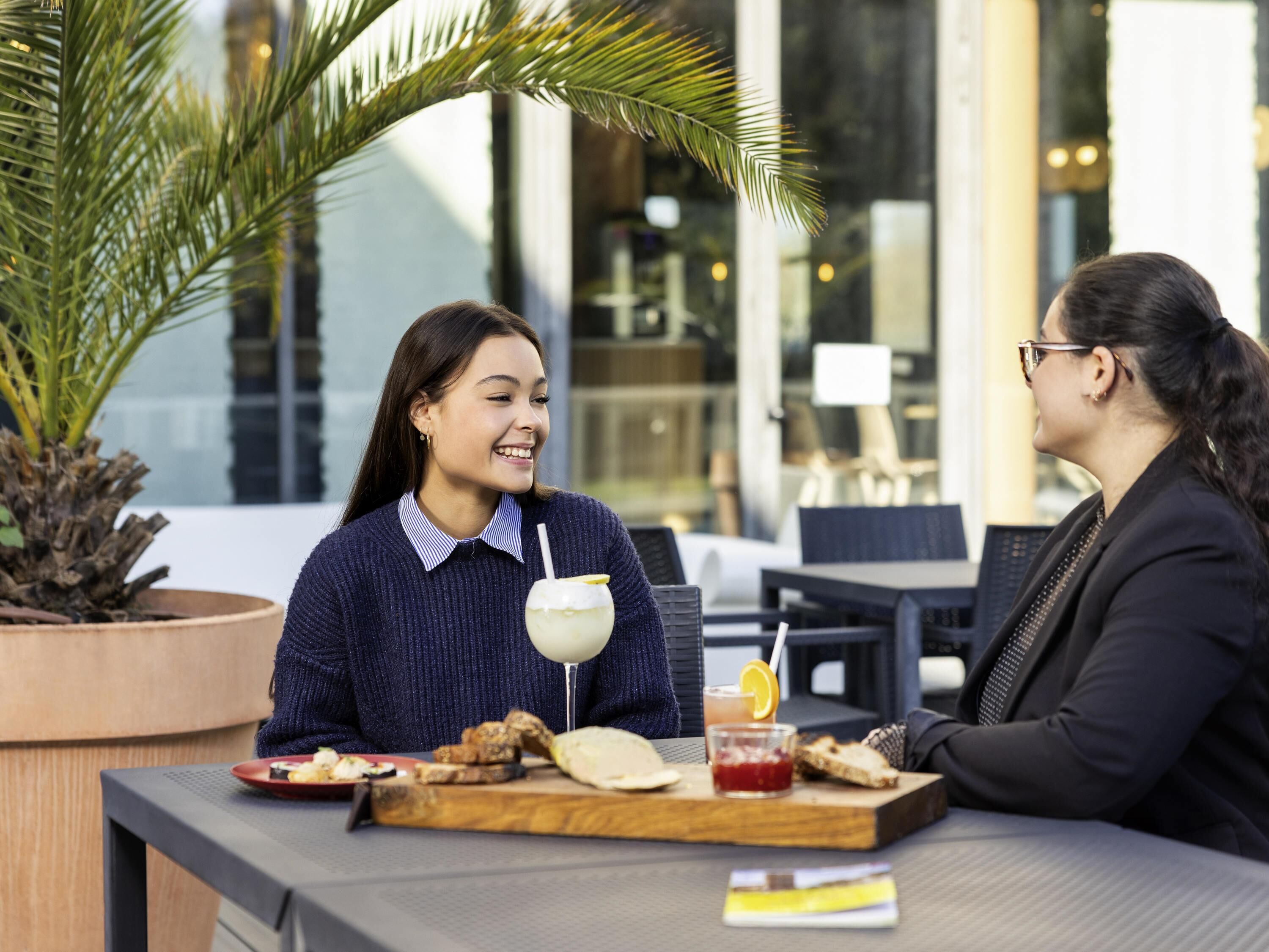 Duas pessoas sentadas em uma mesa ao ar livre, com comida e bebida, uma palmeira e um edifício moderno ao fundo.
