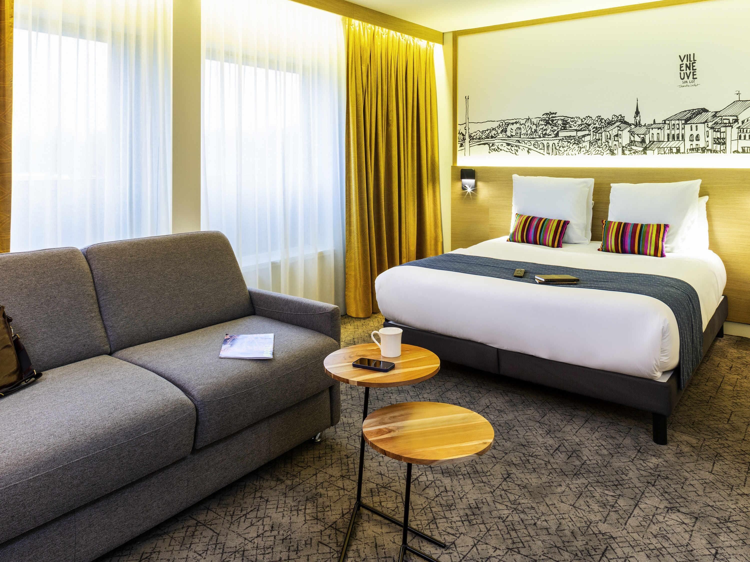 A hotel room with a sofa, a bed, a small table, and a wall with a cityscape mural.