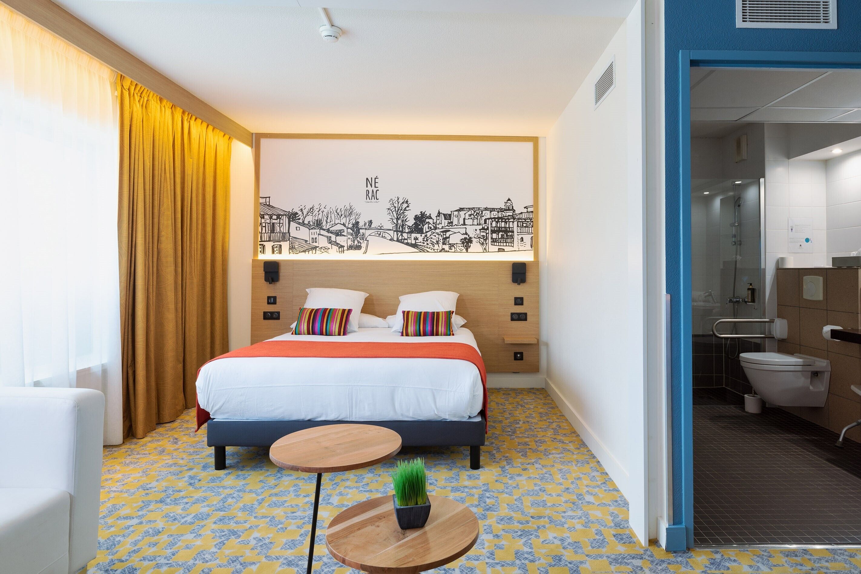 Habitación de hotel con una cama, mesitas de noche, una mesa redonda con una planta, una obra de arte grande colgada en la pared y un baño visible a través de una puerta abierta.