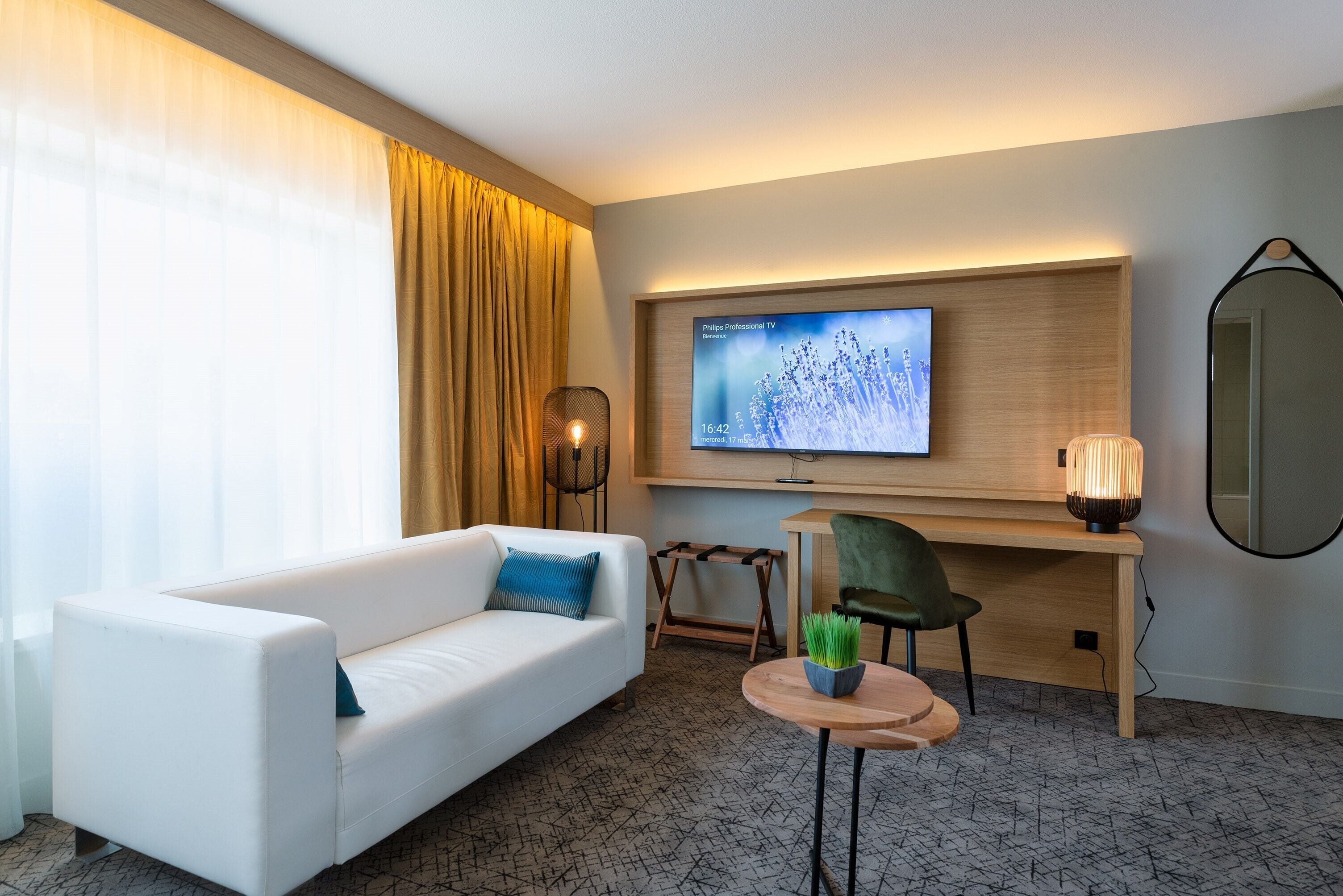 A modern hotel room with a white sofa, a desk with a chair, a television, and a small table with a plant.