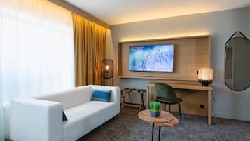 A modern hotel room with a white sofa, a desk with a chair, a television, and a small table with a plant.