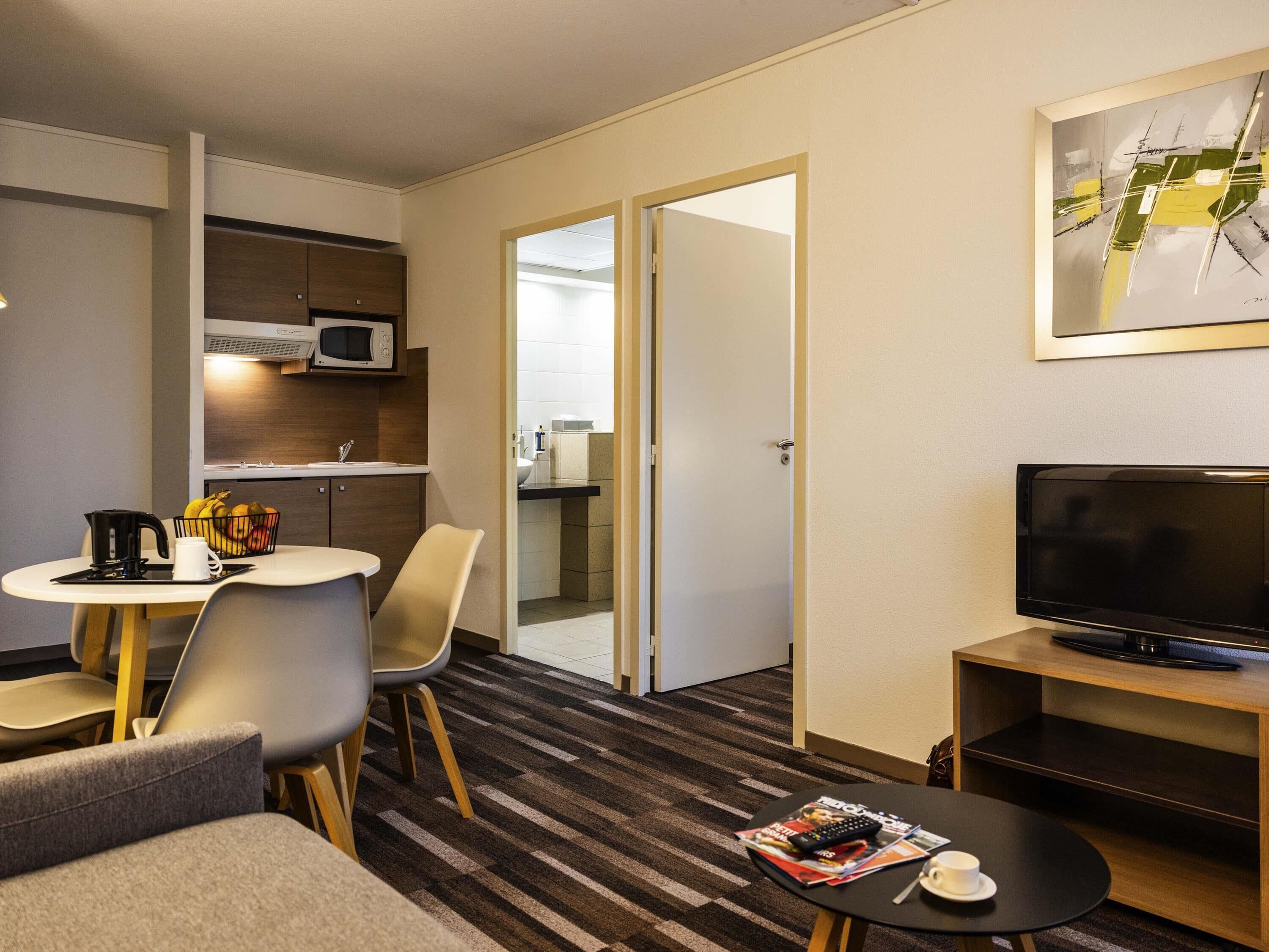 A hotel room with a dining area, kitchenette, a television, and a magazine on the coffee table.