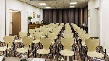 Un salón de conferencias con filas de sillas, un atril y una mesa de recepción con una planta y fotografías enmarcadas.